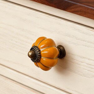 Può includere: Primo piano di un cassetto dell'armadio con un pomello decorativo arancione a forma di zucca. Il pomello ha una base e ferramenta color bronzo. Il cassetto è verniciato color crema con un piano di lavoro in legno scuro.