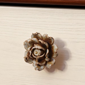 May include: A floral cabinet knob in shades of brown and cream, with a gold central detail. The knob is fixed to a white wooden surface. The knob is likely made of ceramic or porcelain.