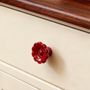 Könnte beinhalten: Nahaufnahme einer cremefarbenen Schublade mit dunkler Holzplatte und einem roten blütenförmigen Knopf. Der Knopf ist mittig auf der Schublade angebracht, und die Holzplatte hat eine satte, dunkelbraune Farbe. Ein geflochtener Korb ist teilweise sichtbar.