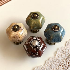 Peut inclure: Quatre boutons de meuble décoratifs beiges, verts, bleus et bruns. Chaque bouton présente un motif tacheté unique et une base métallique. Les boutons hexagonaux ont un élément décoratif central.
