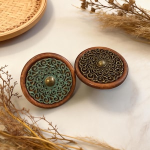Puede incluir: Dos tiradores decorativos de madera para armarios. Cada tirador presenta un diseño circular con una base de madera, una superposición de filigrana metálica detallada y un botón central de color latón. La filigrana tiene una pátina turquesa en un tirador y un acabado bronce en el otro.