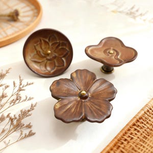 May include: Three bronze-coloured cabinet knobs with floral designs. One is a round knob with a lotus flower, another is a flower-shaped knob, and the third is a cloud-shaped knob. Each knob has a gold-coloured centre.