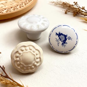 Peut inclure: Trois boutons de meubles décoratifs en céramique. Un est blanc avec un bord festonné, un autre est blanc cassé avec un motif floral, et le troisième est blanc avec un motif floral bleu. Idéal pour les meubles.