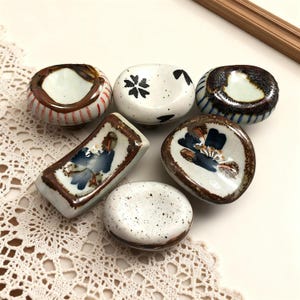 May include: A selection of six small ceramic dishes and holders. The pieces are in various shapes, including round and rectangular, with brown, white, and blue colour schemes. Some have floral or abstract designs, while others have striped or speckled patterns.