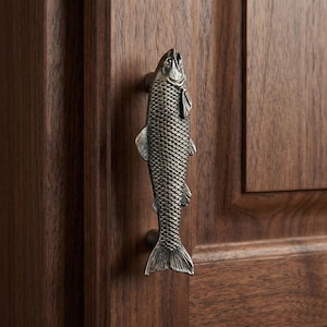 May include: A decorative cabinet pull in the shape of a fish. The pull is made of metal with a textured, scaled surface and is mounted on a dark wood cabinet door. The fish is oriented vertically, with its head at the top.