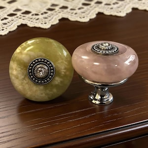 May include: Two decorative cabinet knobs. One is a pale green and white marbled stone with a silver and crystal centre. The other is a pink stone with a silver base and crystal centre. Both are round.