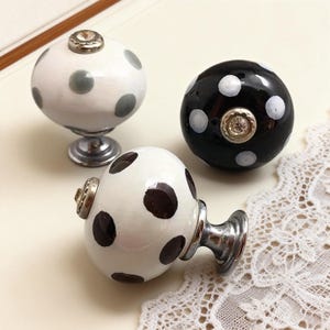 Peut inclure: Trois boutons de meuble décoratifs à pois. L'un est blanc à pois gris, un autre noir à pois blancs, et le troisième blanc à pois marron foncé. Chaque bouton a une base argentée et un dessus décoratif.