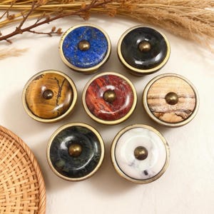 Peut inclure: Ensemble de sept boutons de meubles décoratifs avec des dessus en pierre de différentes couleurs, dont le bleu, le marron et le rouge. Chaque bouton a une base en métal couleur laiton et un accent central en laiton. Les boutons sont disposés sur une surface claire.