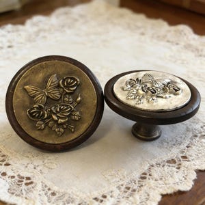 May include: Two decorative cabinet knobs. One knob features a bronze-coloured butterfly and rose design on a dark brown wooden base. The other knob has a silver-coloured butterfly and rose design on a dark brown wooden base.