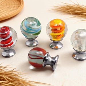 Puede incluir: Un conjunto de pomos decorativos de vidrio con diseños coloridos y arremolinados. Cada pomo presenta un patrón único en tonos rojos, azules, verdes y amarillos, montado sobre una base plateada. Ideal para muebles.
