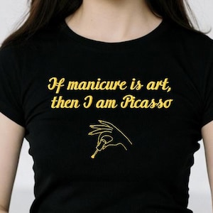 May include: Black t-shirt with gold text that reads "If manicure is art, then I am Picasso." The shirt features a gold line drawing of a hand holding a nail tool. The model is wearing the shirt.