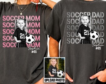 May include: Two charcoal gray t-shirts featuring a custom photo of a young soccer player holding a ball. The left shirt displays pink text reading SOCCER MOM, while the right shirt shows white text reading SOCCER DAD. Both include the name Sophia and #21.