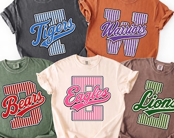 May include: Five short-sleeve crew neck t-shirts in pepper, yam, moss, ivory, and espresso colors. Each shirt features a large block letter with vertical stripes and a script team name overlay: Tigers, Warriors, Bears, Eagles, and Lions.
