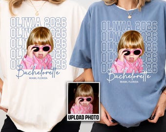 May include: An ivory and a blue jean colored t-shirt, each featuring a graphic print of a child wearing pink sunglasses. The text reads OLIVIA 2026, Bachelorette, and MIAMI, FLORIDA. A small inset box shows the original photo used for the print.