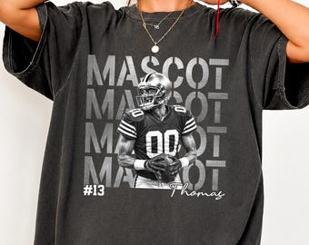May include: A charcoal grey short-sleeve t-shirt featuring a graphic of a football player in a jersey with the number 00. The word MASCOT is repeated in the background, with the text #13 and Thomas printed below the player in white ink.