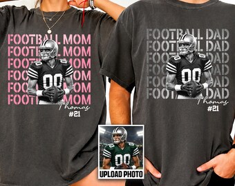 May include: Two charcoal gray t-shirts featuring a graphic of a football player in a helmet and jersey holding a ball. The left shirt has pink text reading FOOTBALL MOM, and the right shirt has white text reading FOOTBALL DAD, both with Thomas #21.