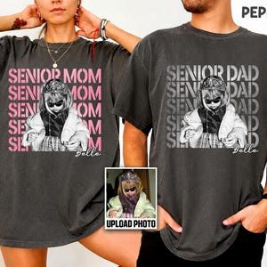 May include: Two charcoal gray t-shirts featuring a graphic print of a child wearing sunglasses and a crown. The left shirt displays the pink text SENIOR MOM, and the right shirt displays the white text SENIOR DAD. Both include the name Belle.