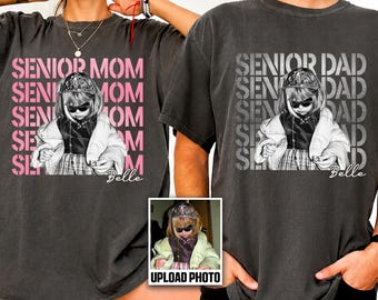 May include: Two charcoal gray t-shirts featuring a graphic print of a child wearing sunglasses and a crown. The left shirt displays the pink text SENIOR MOM, and the right shirt displays the white text SENIOR DAD. Both include the name Belle.