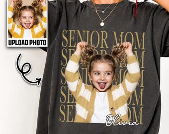 May include: Dark gray t-shirt featuring a photo of a young girl sticking her tongue out, wearing a yellow and white striped cardigan. The shirt has the words "SENIOR MOM" in gold lettering and the name "Olivia" in script.