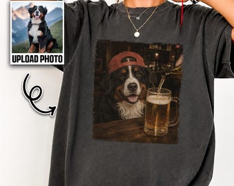 May include: A charcoal gray t-shirt featuring a graphic print of a Bernese Mountain Dog wearing a red baseball cap, sitting at a table with a glass mug of beer being poured. The shirt includes a small inset box showing a dog photo and text.