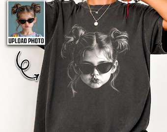 May include: A charcoal gray oversized t-shirt featuring a large, centered black and white graphic print of a young child with hair in two buns, wearing cat-eye sunglasses and making a pouting expression. The word PEPPER is printed in the top right corner.