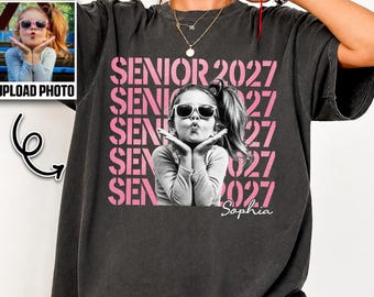 May include: A charcoal gray t-shirt featuring a graphic print of a person wearing sunglasses and blowing a kiss. The text SENIOR 2027 is repeated in pink behind the person, and the name Sophia is written in white script below the graphic.