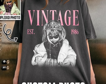 May include: A charcoal gray t-shirt featuring a graphic print of a person wearing a crown and sunglasses, with the text VINTAGE EST. 1986 in pink. The design includes a smaller inset photo of the same person in a light green jacket.