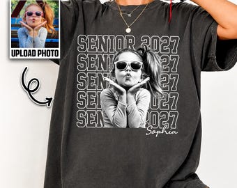 May include: A charcoal gray short-sleeve t-shirt featuring a repeating white graphic print of the text SENIOR 2027 and a black and white photograph of a person wearing sunglasses and blowing a kiss, with the name Sophia printed below the image.