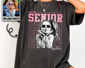 May include: A charcoal gray short-sleeve t-shirt featuring a graphic print of a person wearing sunglasses and blowing a kiss. The design includes the text EST 2027, SENIOR, and the name Sophia in a script font, with a small inset photo box.