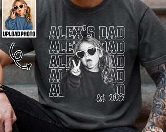 May include: A charcoal gray short-sleeve t-shirt featuring a white graphic print of a child wearing heart-shaped sunglasses and making a peace sign. The text reads ALEX'S DAD repeated in a block font with Est. 2022 printed at the bottom right.