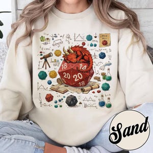 Op de afbeelding: Crèmekleurige sweatshirt met een rode draak die slaapt op een grote, rode twintigzijdige dobbelsteen. Het ontwerp bevat dobbelstenen, wetenschappelijke diagrammen en de tekst "DICE ROLL". Een grillig ontwerp voor fans van fantasiespellen.