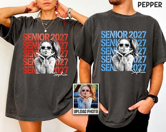 May include: Two charcoal gray t-shirts featuring a black and white photograph of a child wearing sunglasses and blowing a kiss. The design includes the repeated text SENIOR 2027 in red or blue, with the name Sophia written in white script below.