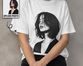 May include: A white short-sleeved t-shirt featuring a large black and white photographic print of a person with a dark bob haircut. The graphic shows the person from the chest up, wearing a dark top. A small inset box shows the original photo.
