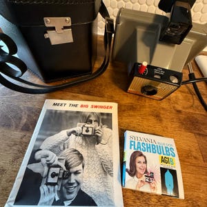 May include: A collection of vintage photography equipment, including a black leather camera case, a gray Polaroid camera, and a flash unit. Also included are two vintage booklets, one titled "Meet the Big Swinger" and the other advertising Sylvania Blue Dot Flashbulbs.