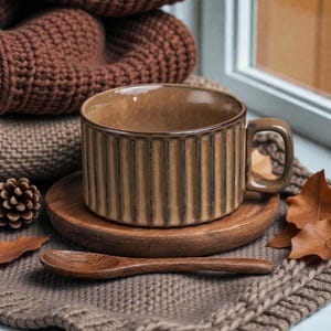 May include: A brown ceramic mug with vertical lines, resting on a wooden saucer. A wooden spoon lies nearby, with a knitted blanket and autumn leaves in the background. The mug has a handle and a textured surface.