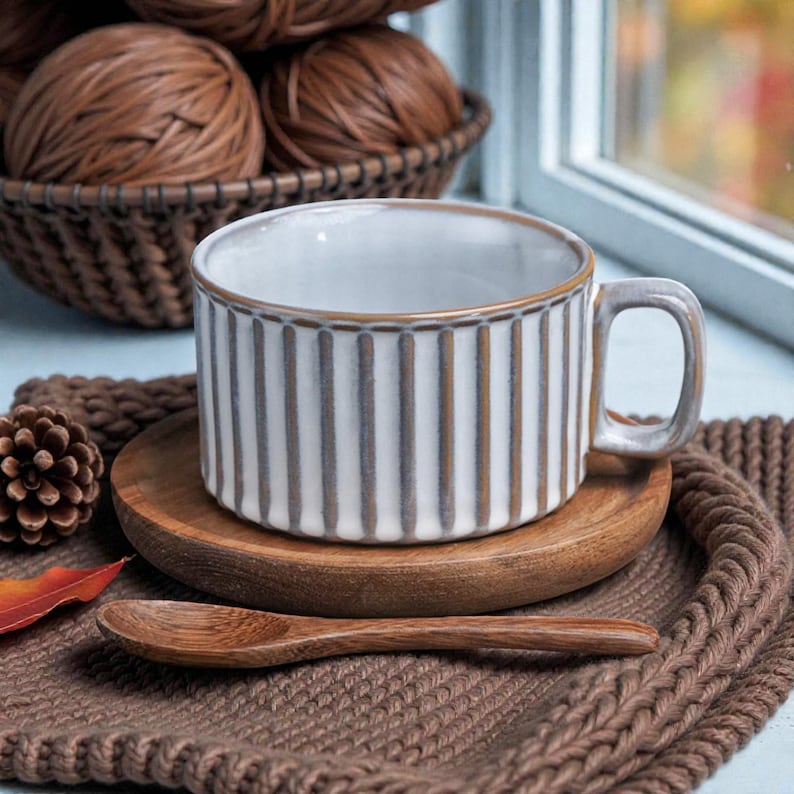 May include: A white and gray striped ceramic mug with a handle, resting on a small wooden plate. A wooden spoon and a brown knitted coaster are in the foreground. A basket of brown yarn balls is in the background.