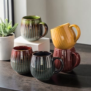 Puede incluir: Un conjunto de seis tazas de cerámica en varios colores, incluyendo verde, amarillo, burdeos y verde azulado oscuro. Cada taza tiene un diseño acanalado y una forma redondeada con un asa. Las tazas están dispuestas sobre una superficie de madera oscura.