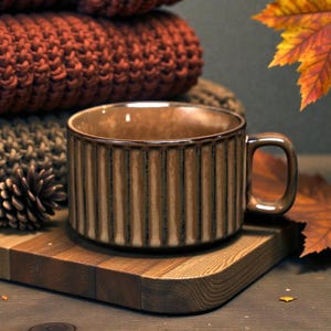 May include: A brown ceramic mug with vertical ribbed detailing sits on a wooden cutting board. The mug has a handle and a warm, earthy tone. A stack of orange knitted items and fall foliage are in the background.