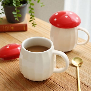 May include: Two white ceramic mugs with a faceted, rounded body and a handle. One mug is filled with a brown liquid and sits next to its separate red lid with white polka dots. The other mug is covered by an identical red and white mushroom-style lid.