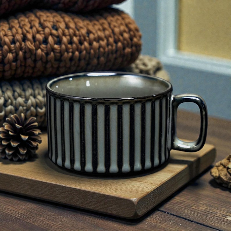 May include: A ceramic mug with vertical black and white stripes, a rounded handle, and a dark rim. The mug sits on a small wooden cutting board. A stack of brown knitted items and pine cones are in the background.
