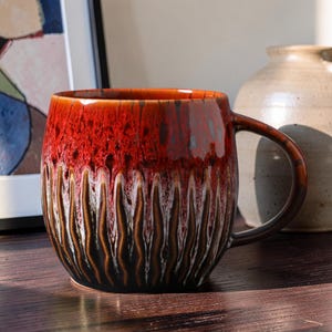 May include: A ceramic mug with a glossy, textured finish. The mug has a gradient of red, brown, and black hues. The design includes vertical, wavy lines. The handle is brown. The mug is on a wooden surface.