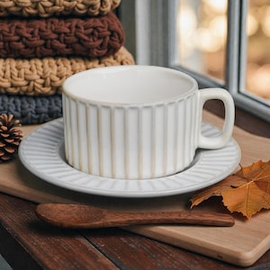 May include: A white ceramic teacup and saucer set with vertical ribbed detailing. A wooden spoon rests on a wooden cutting board. A stack of knitted items in brown, tan, and grey are in the background.