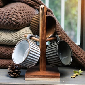 May include: A wooden mug tree holds three ceramic mugs with striped and solid designs. The mugs are in shades of white, gray, and brown. A stack of brown knitted blankets and a pinecone are in the background.