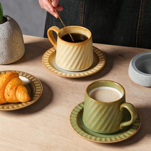 May include: Two ceramic coffee cups with matching saucers. One cup is beige with dark coffee, the other is green with a creamy beverage. Both cups have a ridged design. A croissant sits on a plate nearby.