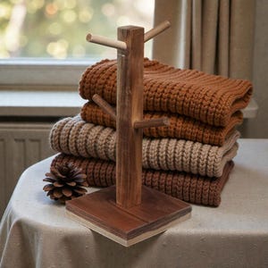 May include: A wooden mug tree with a square base and multiple pegs for hanging mugs. A stack of folded, knitted blankets in shades of brown and tan sits behind the mug tree. A pinecone rests on the table in front of the tree.