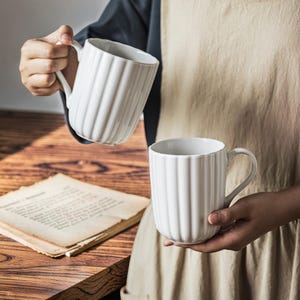 May include: Two white ceramic mugs with vertical ridged detailing. The mugs have a curved handle and a rounded base. One mug is held in the air, while the other is held in a person's hands. The mugs are on a wooden surface.