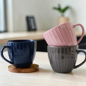 Puede incluir: Tres tazas de cerámica en azul marino, rosa empolvado y gris, apiladas y dispuestas sobre una superficie de madera. Cada taza tiene un diseño acanalado y un asa curva. Una taza azul marino está sobre un posavasos de madera.