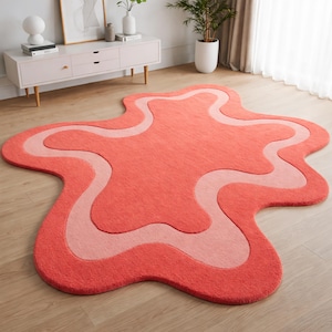 May include: A coral-colored, abstract-shaped rug with a lighter pink wavy design. The rug is placed on a wooden floor, adding a pop of color to the room. The rug's unique shape and color palette make it a statement piece.