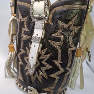 Puede incluir: Un bolso de mano de cuero negro hecho de una bota de vaquero, con costuras en forma de estrella y un cinturón de cuero blanco con hebilla plateada. El bolso tiene flecos y una correa para el hombro.