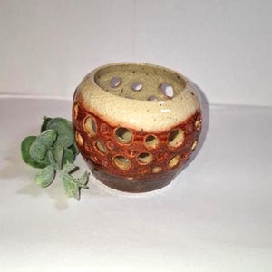 Puede incluir: Un portavelas de cerámica con un diseño único. El soporte es redondo con una parte superior color crema y una base marrón rojizo. Cuenta con numerosos recortes circulares, creando una pieza decorativa y funcional. Una ramita de follaje verde está al lado del portavelas.